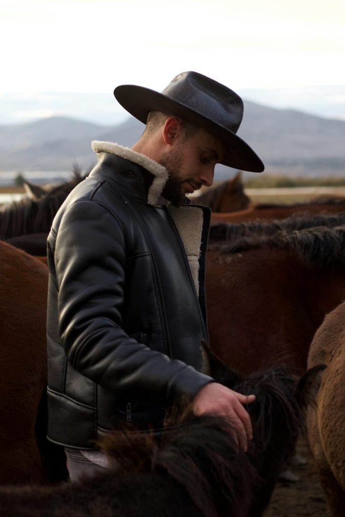 pexels-photo-14174936-14174936 Man in a cowboy hat and leather jacket standing among horses on a ranch.