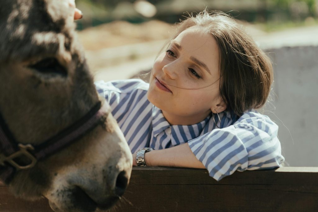 A young woman in a striped shirt affectionately interacts with a donkey outdoors on a sunny day.